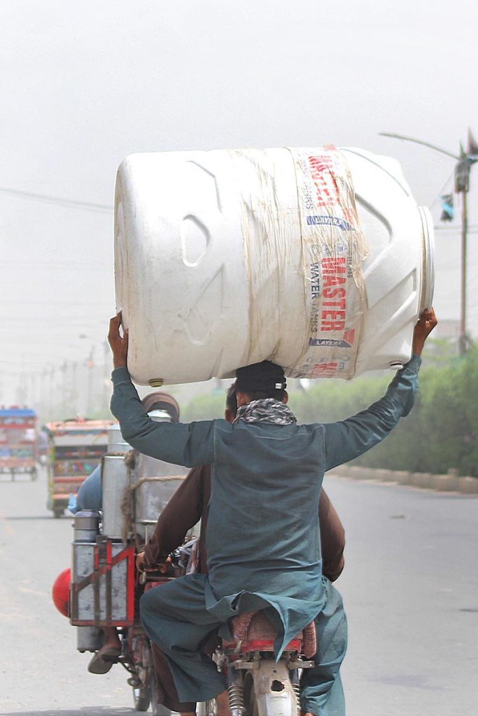 A pillion rider carrying water tank on his head heading towards his destination