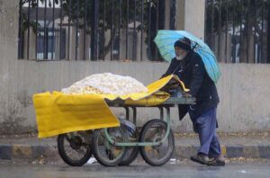 An aged vendor on the way under the cover of an umbrella to protect him from the first rain of the season.