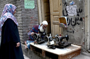 An aged man repairing the cloth sewing machine of a woman at his setup in a street of walled city